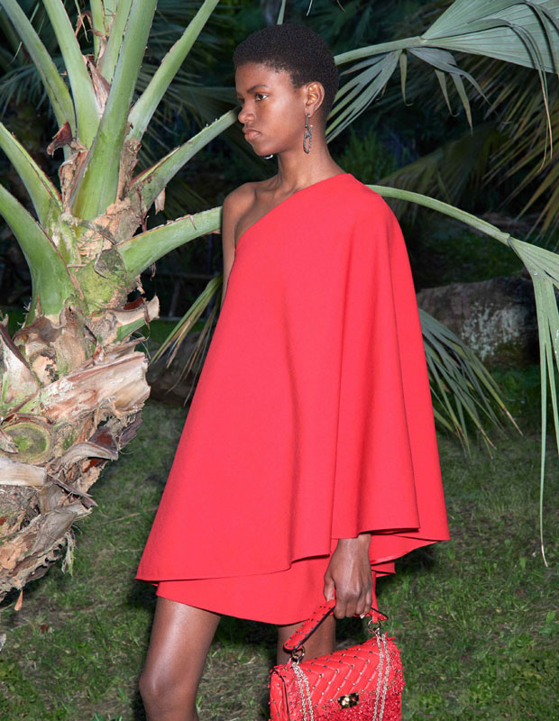 A model wears an asymmetrical red dress and stands under a palm tree