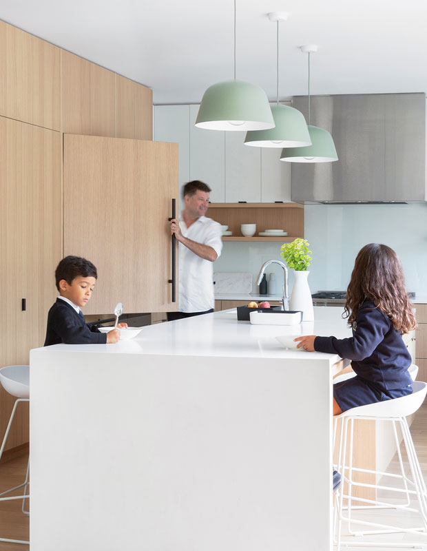 Family sitting in a minimalist kitchen with seafoam green pendant lights on the ceiling and pale wooden cabinetry.