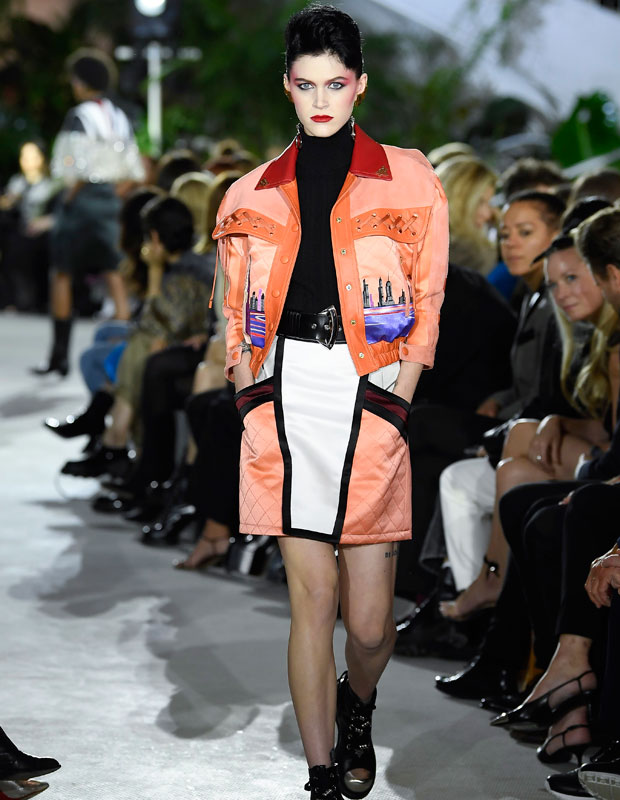 A model walks on the runway wearing a peach colored skirt and jacket with red and black accents
