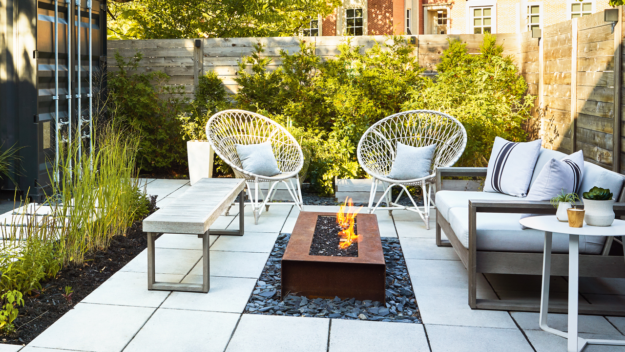 A backyard patio with a square metal fire pit in the center, two white chairs on the side, a wooden bench, and green plants all around
