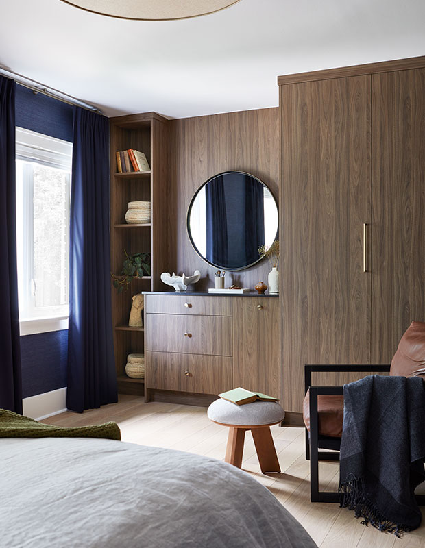 a bedroom with a large walnut closet and vanity set with a large round MCM mirror, and textured navy wallpaper with matching curtains. little italy home renovation