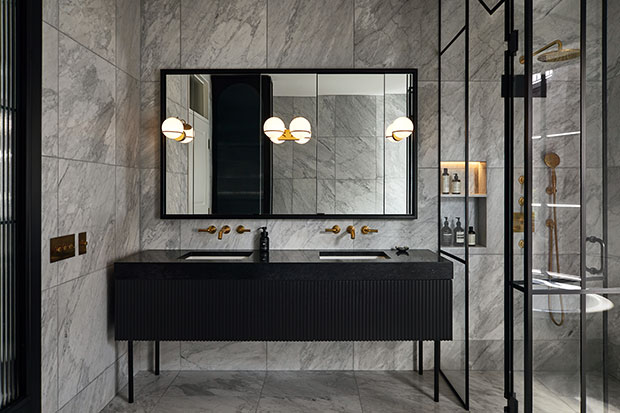 A wide shot of a slick, modern bathroom. Large grey square marble tiles. line the walls. Black metal and glass create a modern shower area, and a black clawfoot bathtub with white interior. A modern spherical light and gold hardware accent the room. A large black double vanity sits at the wall, under a large mirror with more spherical sconces attached.