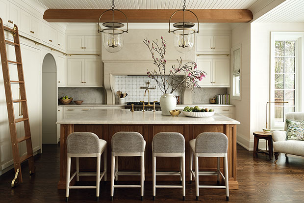 A modern farmhouse style kitchen with exposed wood beams, cream cabinets, a large quartz island with a wood base. on the left wall, an arched doorway leads to a pantry, and a wooden ladder leads to the high cabinets. four chairs sit at the island. two minimal yet-stylish light fixtures hang from the ceiling in brass and glass.