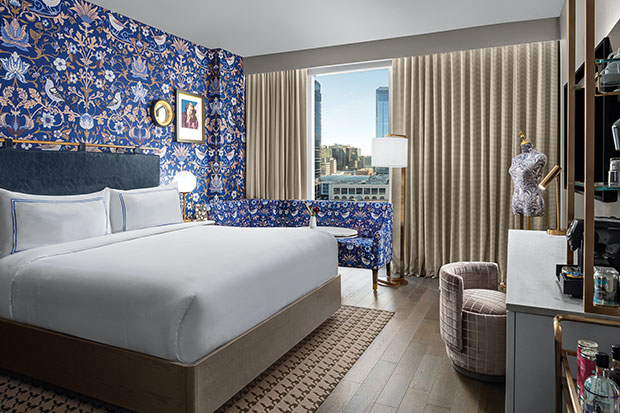 A wide shot of a pretty hotel room, in which the walls are lined with bold floral wallpaper, which is the same print as the couch, and a large bed sits against the wall. A large window looks out on the Calgary skyline, and a large desk/vanity and dress form sit opposite the bed. calgary travel guide