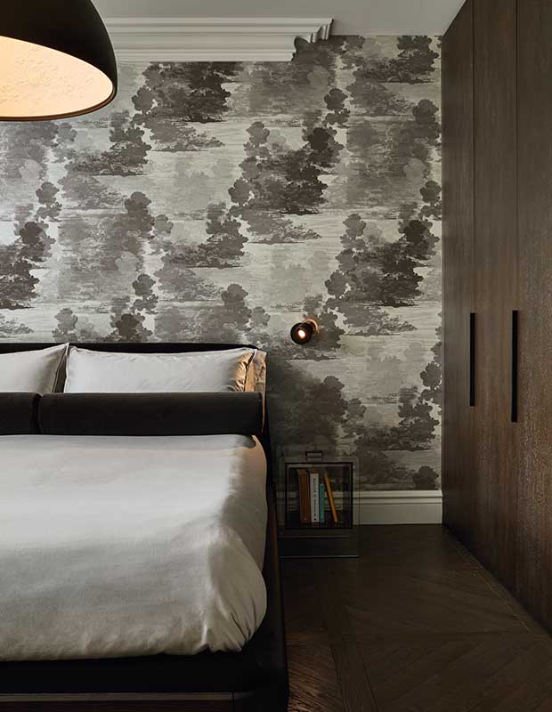 A cropped portrait of the principal bedroom, which has grey toned abstract cloud patterned wallpaper, a large simple bed with grey linens, a very large rounded ceiling light, and a small, minimal bedside table with a few books in it. On the right wall is a large modern dark wood closet.