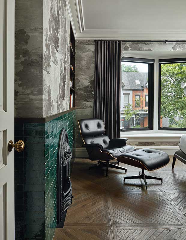 A portrait of half of a principal bedroom, which has printed grey abstract cloud wallpaper, A black iron fireplace with jade green tiling, and large floor-to-ceiling windows. A black leather Eames chair and ottoman sit beside the fireplace.