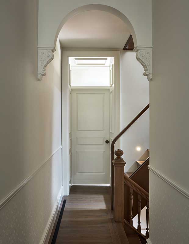 A simple back entryway with white walls and a large window above the door. Dark wooden stair bannisters are visible to the right going down, while a sleek modern glass railing with black metal leads upwards. An archway connecting from one wall to the other stands out as the main design feature.