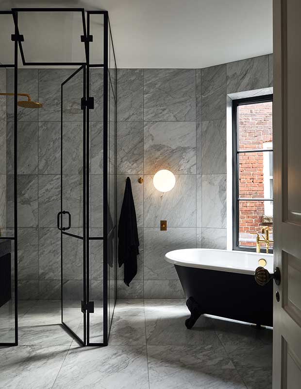 A vertical shot of a principal ensuite with large grey square marble tiled walls. Black metal and glass create a modern shower area, and a black clawfoot bathtub with white interior. A modern spherical light and gold hardware accent the room.