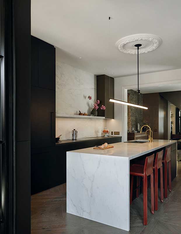 A modern kitchen with black cubic cabinets, a white marble island that matches the backsplash. Deep red metal bar chairs sit under the island, and a long exposed bulb light fixture hangs above it.