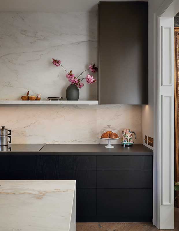 Matching modern and minimal black counter tops and cabinets stand out against the white marble backsplash of this kitchen. a vase of minimal pink flowers sits on a shelf above the counter. A moka pot sits on the sleek stove.
