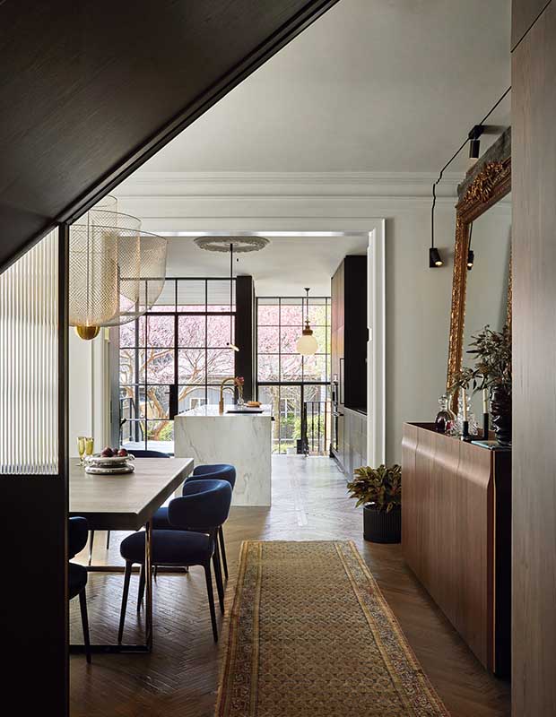 A vertical shot of the dining room, which features a long runner rug, a large wooden dining room table with modern blue velvet chairs, and further, the kitchen can be seen, with a white marble island, and floor-to-ceiling windows. funky lighting hangs above the table and the island, and the walls are painted white which contrasts the dark flooring and furniture.