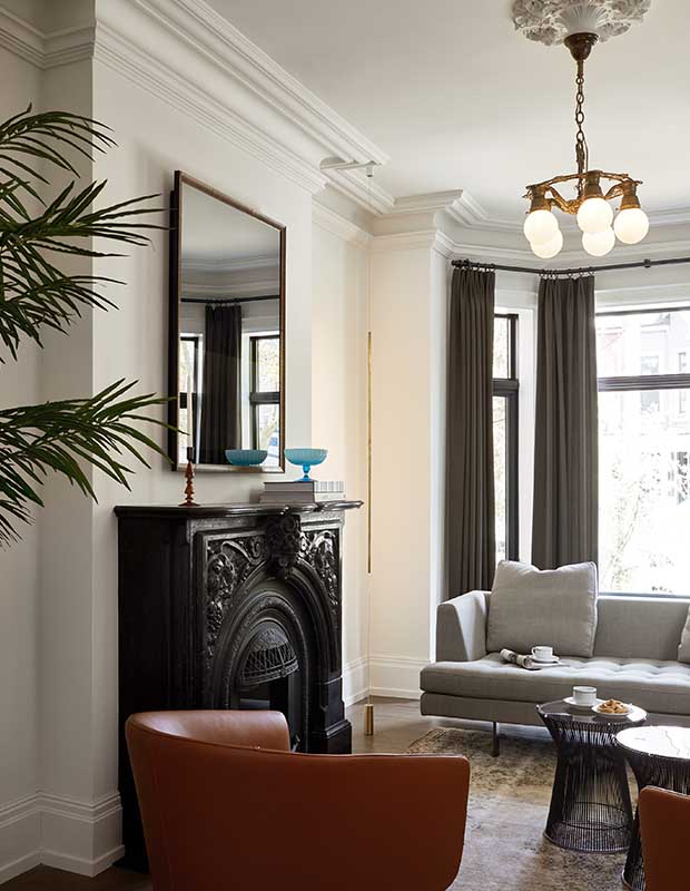 A vertical image of a living room with a black carved fireplace, modern grey sofa, deep dusty orange rounded leather chairs, a tall window letting in natural light, illuminating the classic molding on the ceiling and at the light fixture, which is a vintage gold with modern rounded bulbs.