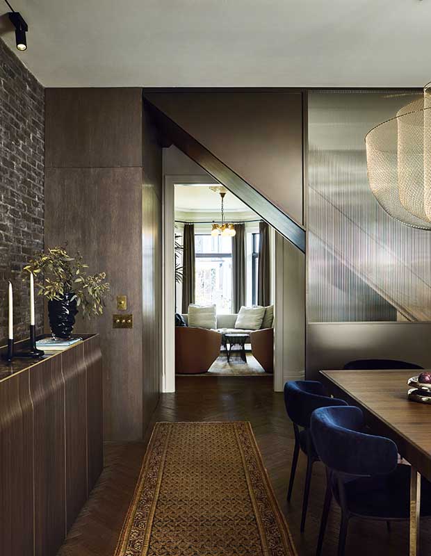 a vertical shot of a dark and moody dining room with a textured glass dividing wall, dark wood floors and walls (with an exposed brick accent wall) and dark blue velvet chairs at the large wooden dining room table. A Persian runner rug leads to a hallway, where the living room is visible beyond.