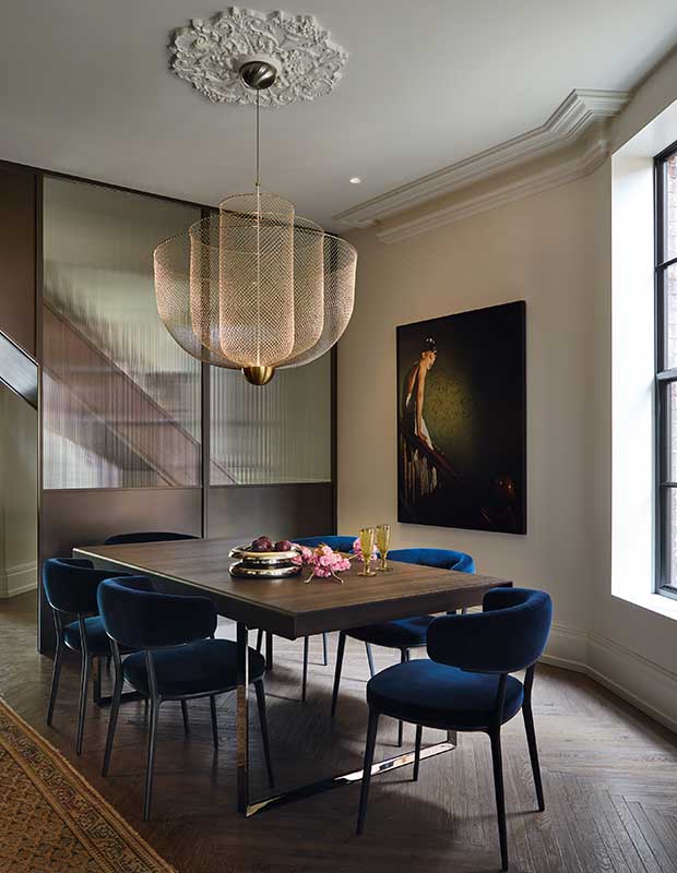 A vertical shot of a dark dining room with a large modern pendant light that resembles wire netting, attached to the ceiling with sculpted molded detailing on the ceiling. A half wood half glass dividing wall separates the dining room from the hallway. A large painting of a woman on stairs hangs on the wall. the floor is dark hardwood.