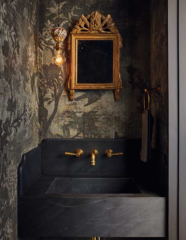 A dark luxurious bathroom with floral wallpaper in a textured black and grey. An ornate gold mirror hangs with a funky exposed hanging lightbulb. a black modern marble sink with gold hardware sits below the mirror.