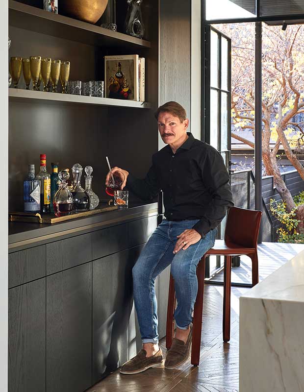 A man (keith) sits beside a minimal modern bar, made from dark wood. various bottles and glasses line the shelves, many of them in cohesive gold tones.