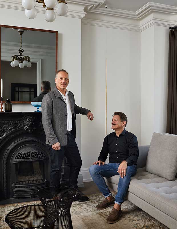 Two men are in the living room, one stands, while the other sits on the grey couch. Behind them is the black carved fireplace and mirror/tv, in which the pendant light reflects. An old Persian rug lines the floor, and on top of it sit two knee-height black wire sculptures.