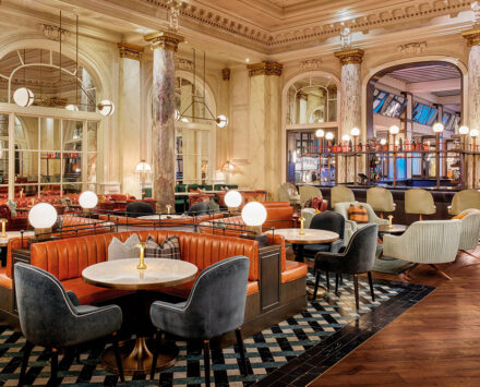 wide shot of a luxurious restaurant dining room with deep orange leather booths and plush suede chairs. The walls and columns are are white marble bathed in soft white light emitted from modern orb lighting fixtures. The floor is an intricate diamond patterned tile.