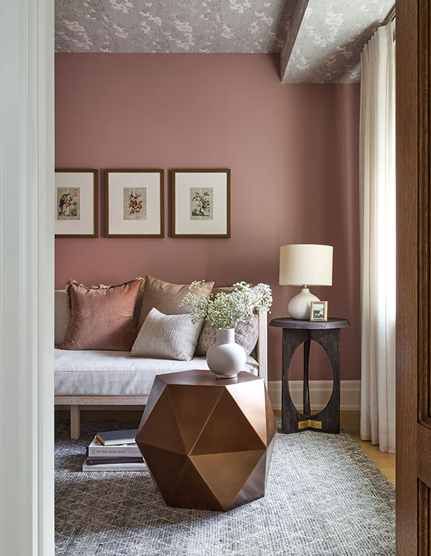 A modern sitting room with dusty pink walls and matching throw pillows. Rounded decor such as lamps and vases, contrast with the angular rose gold side table. a plush grey patterned rug is what the light-wood sofa sits upon. modern heritage townhouse