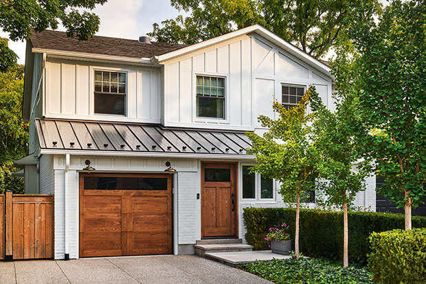 A white home with features that resemble a barn such as wooden paneling and mid-tone barn-style doors for the garage and front door, and a metal roof. the home is surrounded by large green trees. modern heritage townhouse