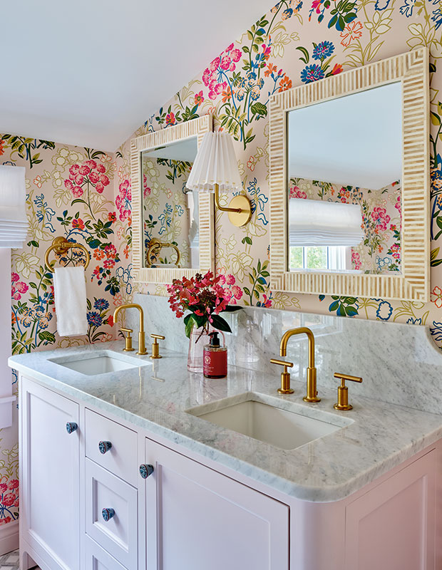 A brightly coloured bathroom featuring a pale pink double vanity with marble countertops, bright floral wallpaper with pink hues, and a retro wall sconce with a white, structured lampshade. old nova scotia home