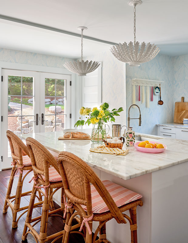 A bright updated kitchen with coral and pale-blue accents, rattan chairs at the island, fresh flowers and fruit on the counter, and unique white plant pendant lighting. Glass French doors lead outside and let lots of natural light in. old nova scotia home