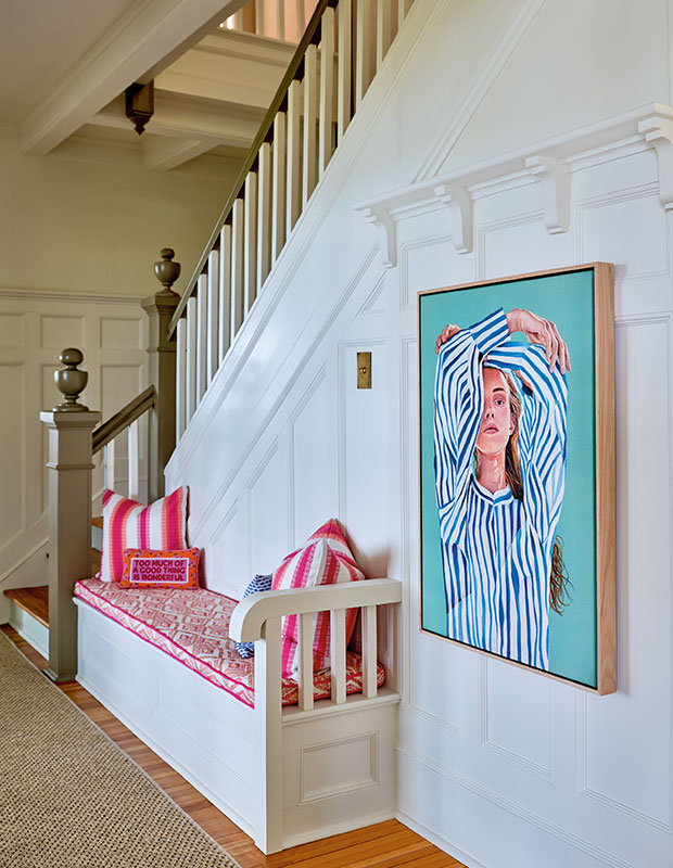 A portrait of a bright hallway with a large white staircase adorned with a painted portrait of a white woman in a striped shirt in front of a bright turquoise background, and a small bench with bright magenta cushions. old nova scotia home