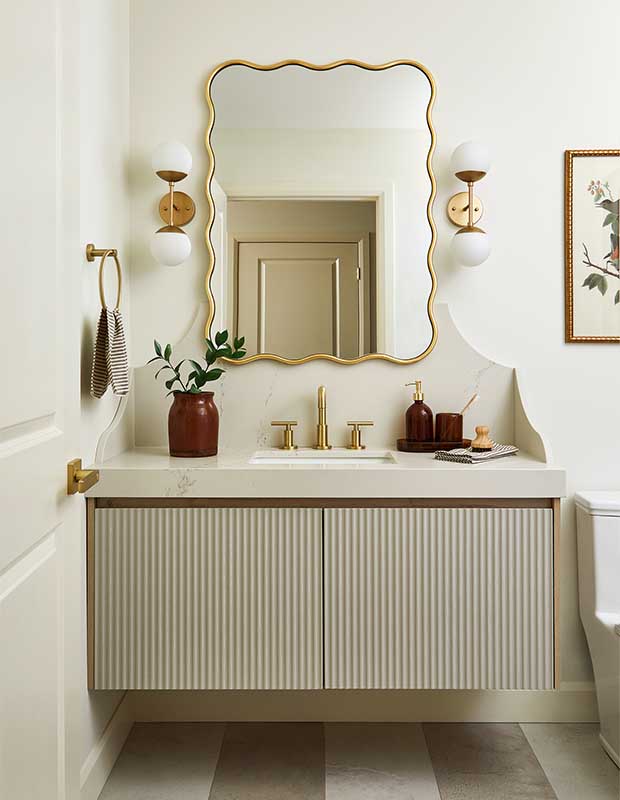 A bathroom accented with gold design elements including mirror, handles, tap, and towel holder. A textured striped cabinet below the sink counter in marble. Matching burgundy vase and soap holder tie the space together with a colour pallet of burgundy, gold, white, and beige. Morrisburg budget reno. stylish budget reno