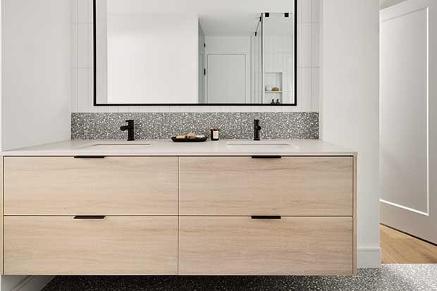 A light wood double vanity with a short grey backsplash that cuts off to a large rectangular mirror in this white-walled bathroom. english cottage-style house