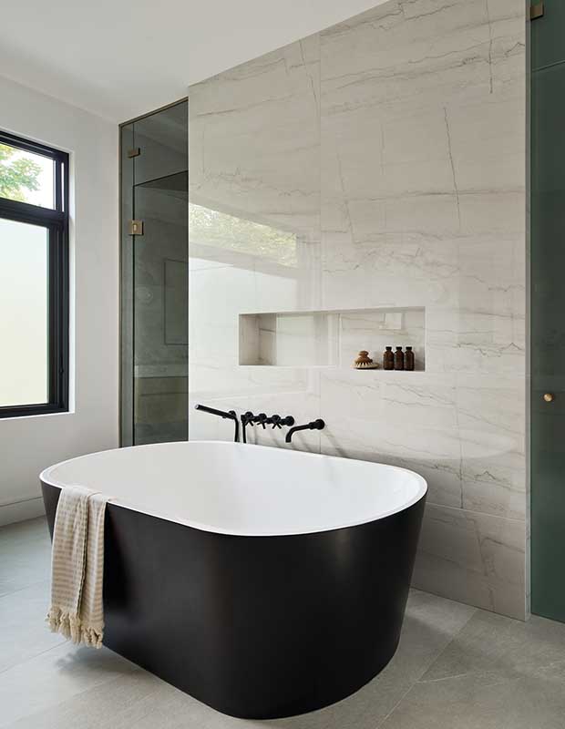 The ensuite bathroom contains a large soaker tub with dark grey lining on the white porcelain bathtub. Floor-to-ceiling grey marble is surrounds the tub. english cottage-style house