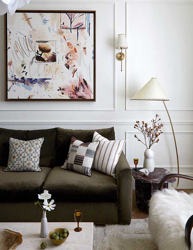 A white living room with a brown couch and patterned throw cushions. An abstract painting sits on the wall, and flowers in vases on both tables. Two lamps with golden accents tie in with other golden accents. notting hill flat