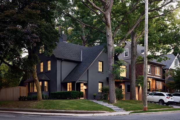 A matte black home sits on the corner of a suburb street surrounded by old trees. english cottage-style house