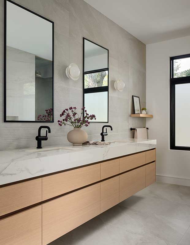 A clean modern bathroom with grey marble tiles that flow from ceiling to floor. a light wood cabinet topped with marble countertops in a double vanity. Matte black hardware ties the room together. english cottage-style house