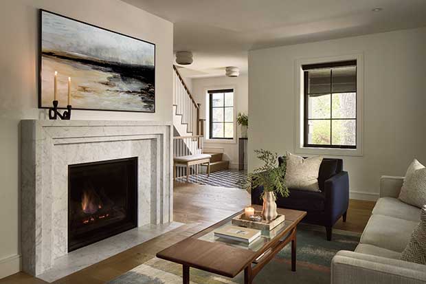 A modern living room with classic elements including a marble fireplace, cool navy sitting chair, grey couch, and a painting of a beach. A staircase and entryway is seen in the distance. english cottage-style house