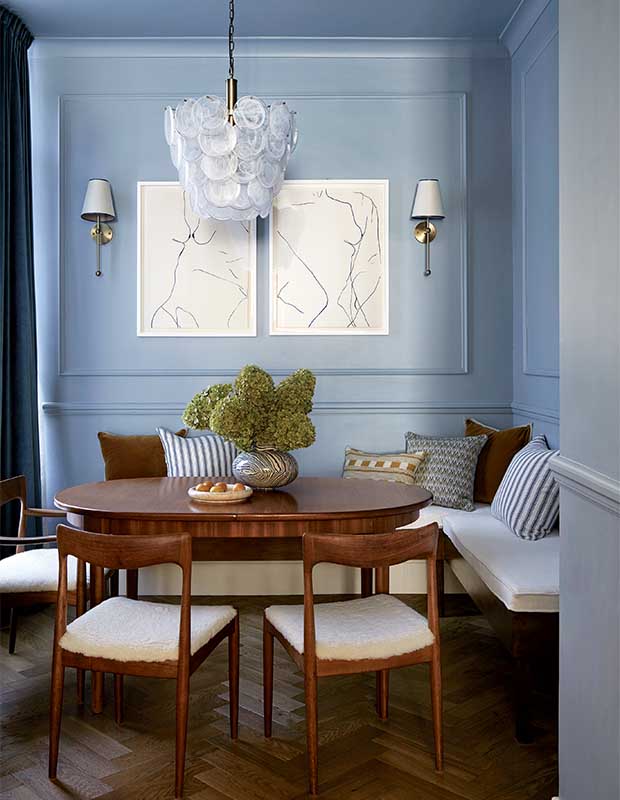 Portrait of a dining room with muted blue walls, medium wood table and chairs, a bench on the far end with striped cushions. a unique pendant light and abstract white art. A small bonsai tree sits in the middle of the table. notting hill flat
