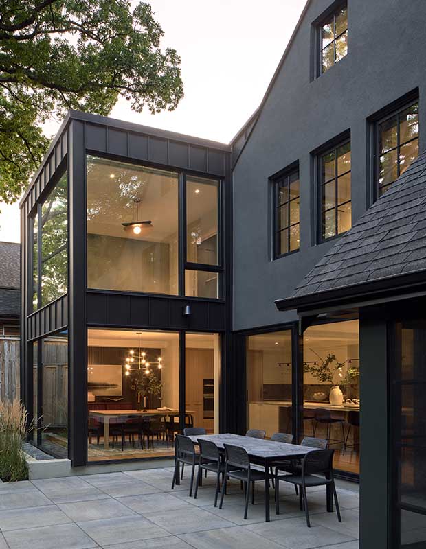 A shot of the back of the home from the backyard. Floor-to-ceiling windows allow views of the warmly-lit home. The exterior is matte black to match the patio dining furniture. english cottage-style house