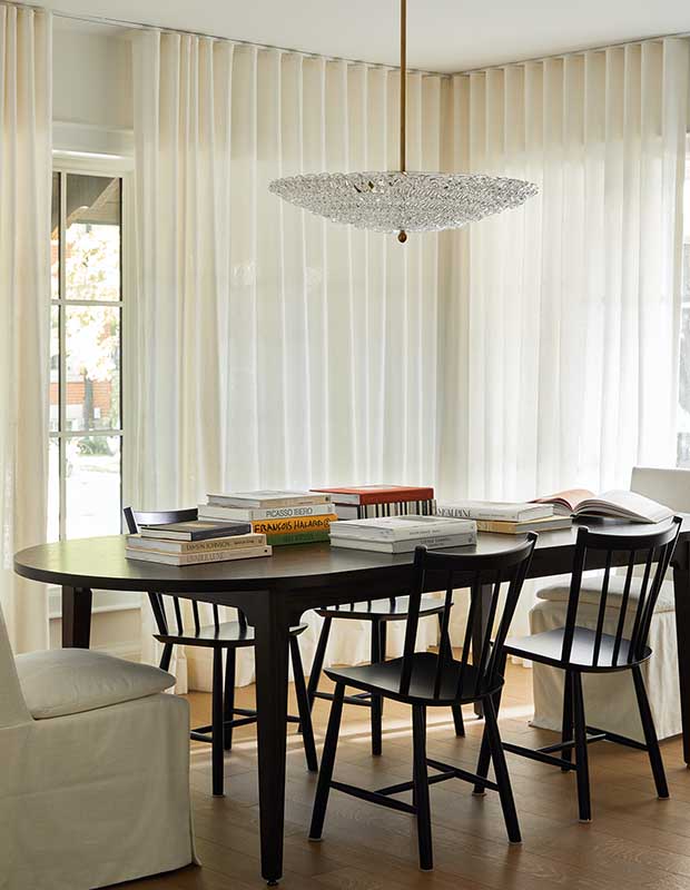 An all white living room with dark-wood dining set. white curtains allow natural light in, a large crystal pendant light hangs above the table. serene city retreat
