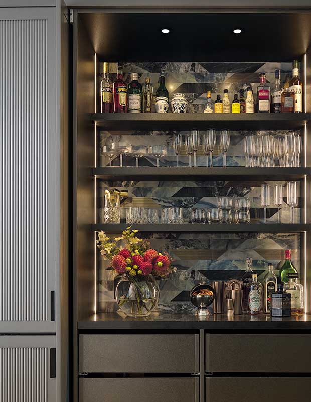 A close up of the bar, which has a tile blackspash, many different types of glasses, and bottles of alcohol. english cottage-style house