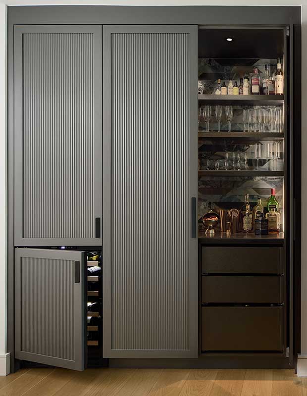 A black bar with tall cabinets with reeded wood doors. an open section displays glasses, bottles, and drawers. english cottage-style house