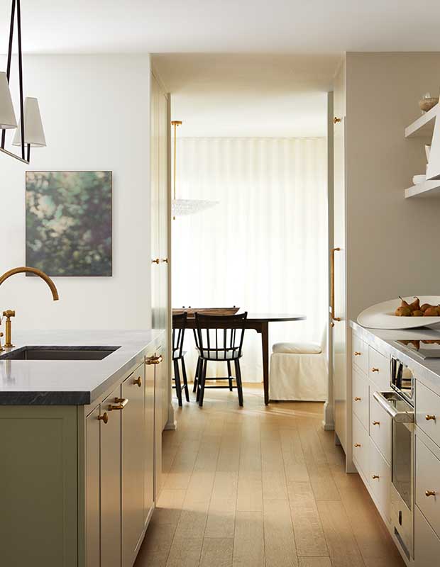 A light kitchen with muted green details, brass hardware, and white walls and accents. In the distance, we see the dining room. serene city retreat