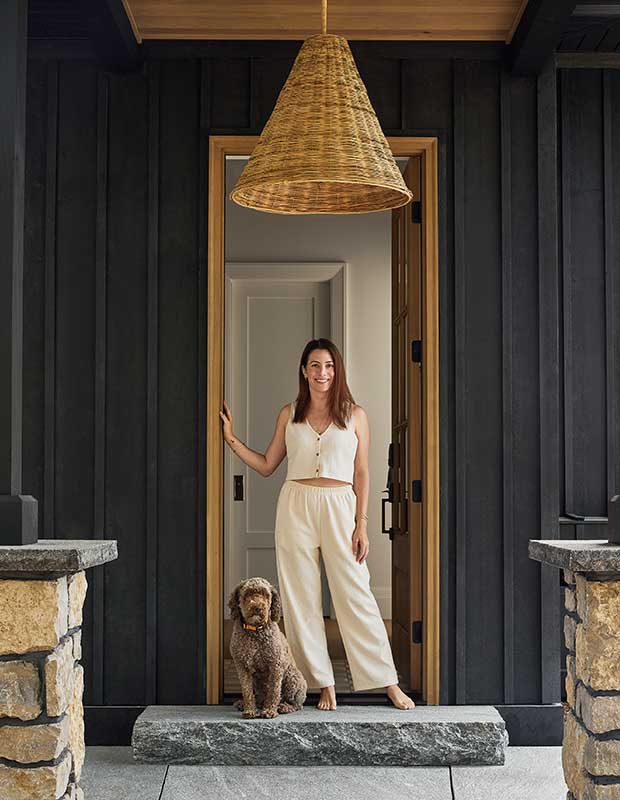 Designer Parris McKenna, a caucasian woman with brown hair, stands in the entryway to a bunkie with black metal walls, a wicker lamp, and a fluffy brown dog sitting next to her. serene muskoka bunkie