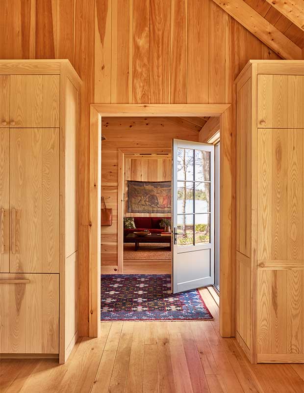 A pine walled entryway is seen with a rustic red and blue carpet, the living room is seen through a doorway. wood-clad family cottage