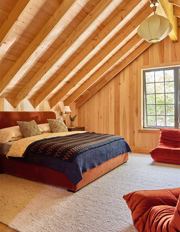 A pine walled room with a slanted ceiling and visible beams. Rusty red bean bag chairs sit on the floor, a knitted blanked sits atop the bed spread. wood-clad family cottage