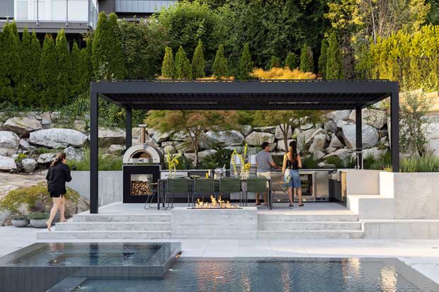A pergola with open sides sits up steps just off the side of a pool, with a dining table and outdoor oven. unique backyard pools