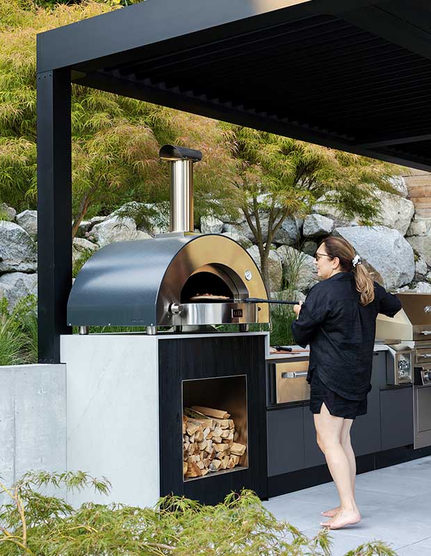 A woman stands at an outdoor pizza oven under a pergola. unique backyard pools