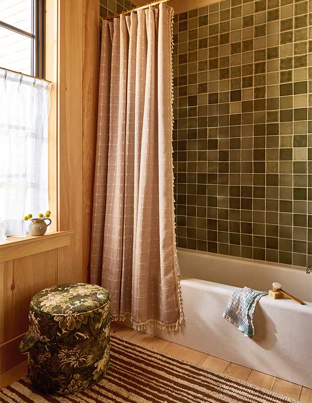 Green tile lines the walls of this bathroom which has a classic white tub, a light pink fringed shower curtain, and a striped rug. wood-clad family cottage