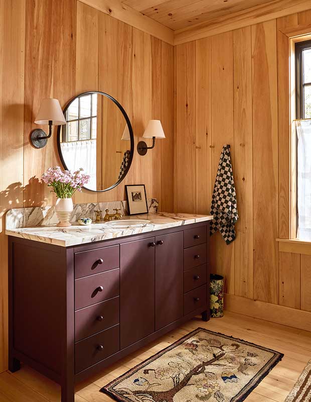 A maroon vanity with marble counters sits in this pine-walled bathroom. A round mirror, wall sconces, and small floral carpet.wood-clad family cottage
