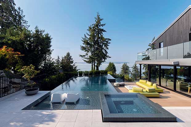 A scenic view of a backyard with a long, infinity style pool with a white deck and large white chairs in the water. The ocean is seen in the background. A large house sits to the right of the pool. unique backyard pools
