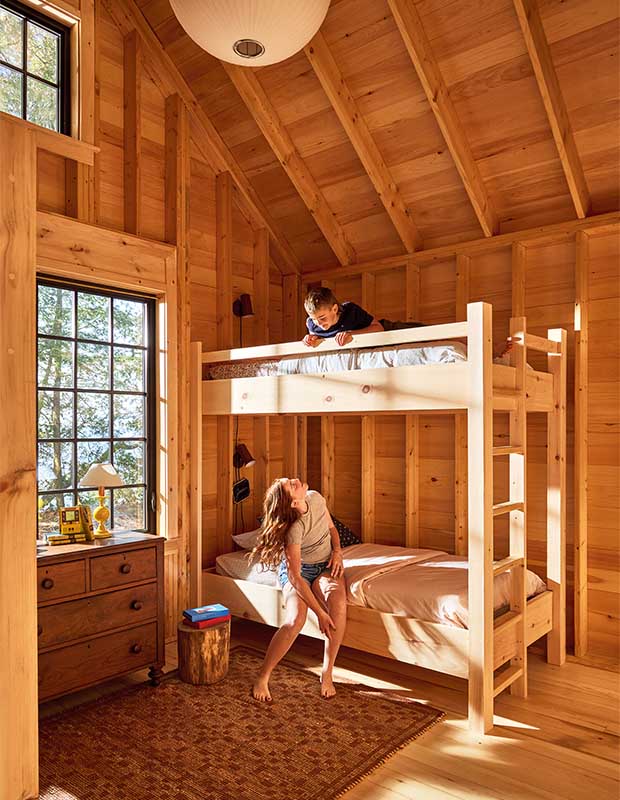 A boy and a girl sit in a pine-walled bedroom on a set of bunk beds, the ceiling is arched, and two windows let in natural light. a patterned red rug sits on the floor. wood-clad family cottage