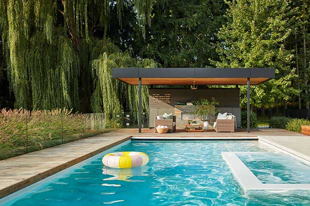 A large pool with a black rimmed pergola at the edge including an outdoor fireplace and wicker chairs. A floaty floats in the blue water. unique backyard pools